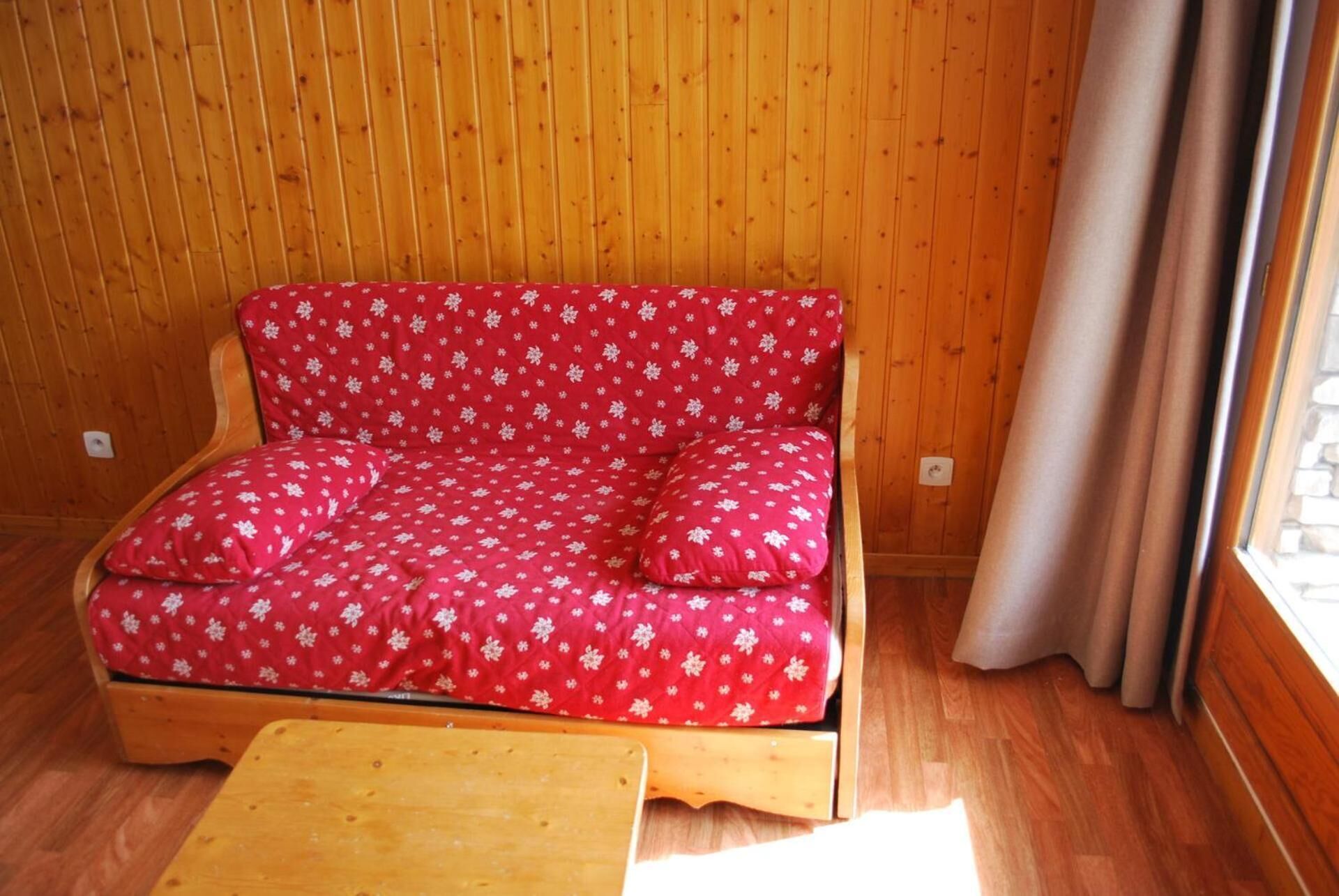 A small wooden room with a red floral sofa and a wooden table.