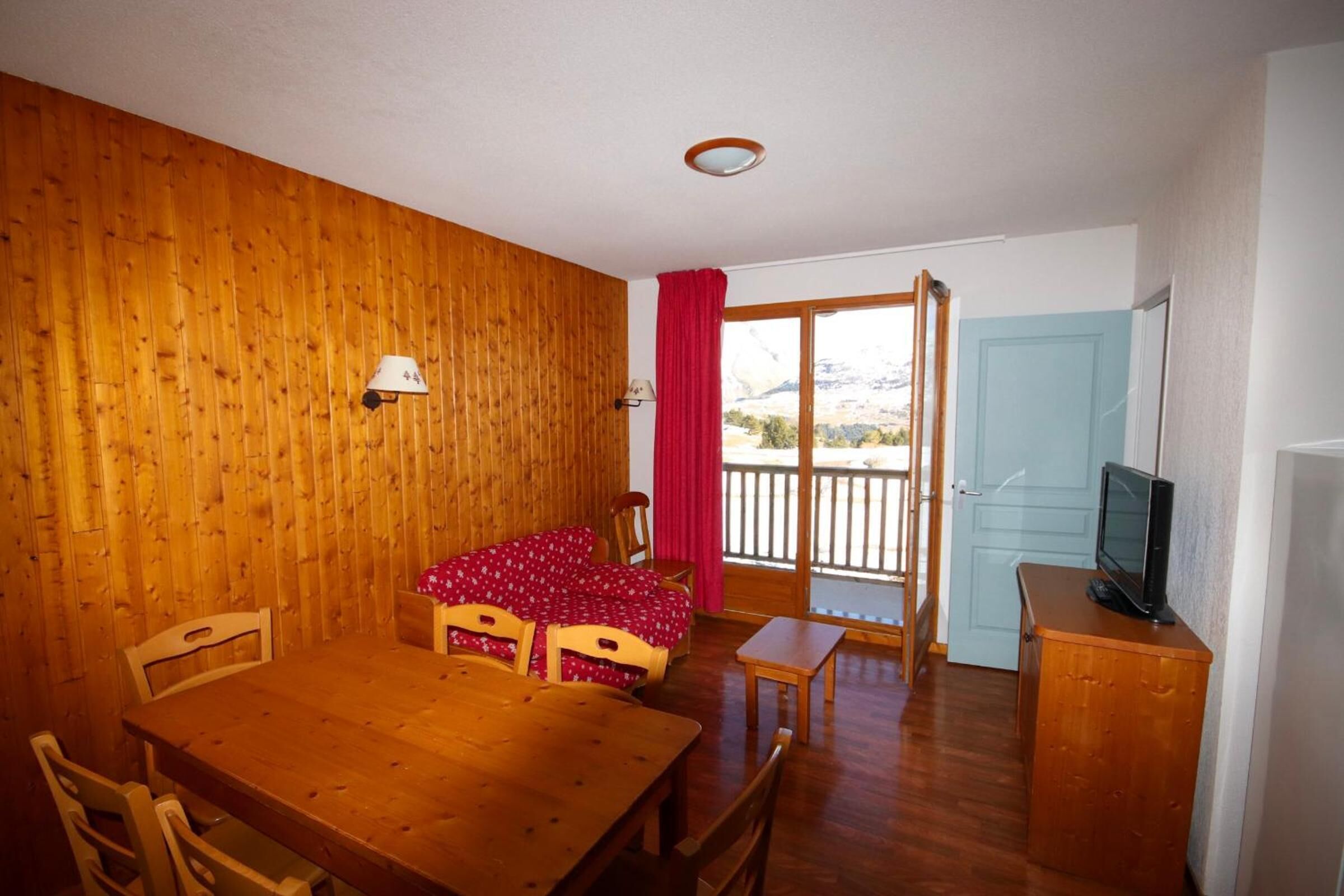 A wooden-paneled room with a dining table, chairs, a small TV on a wooden cabinet, and a red curtain.