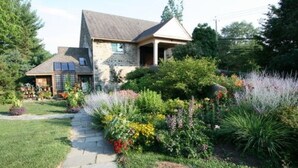 A stone house with a sloped roof, a covered porch, and a garden with various plants and flowers. - Alpenhof Bed and Breakfast (Media)