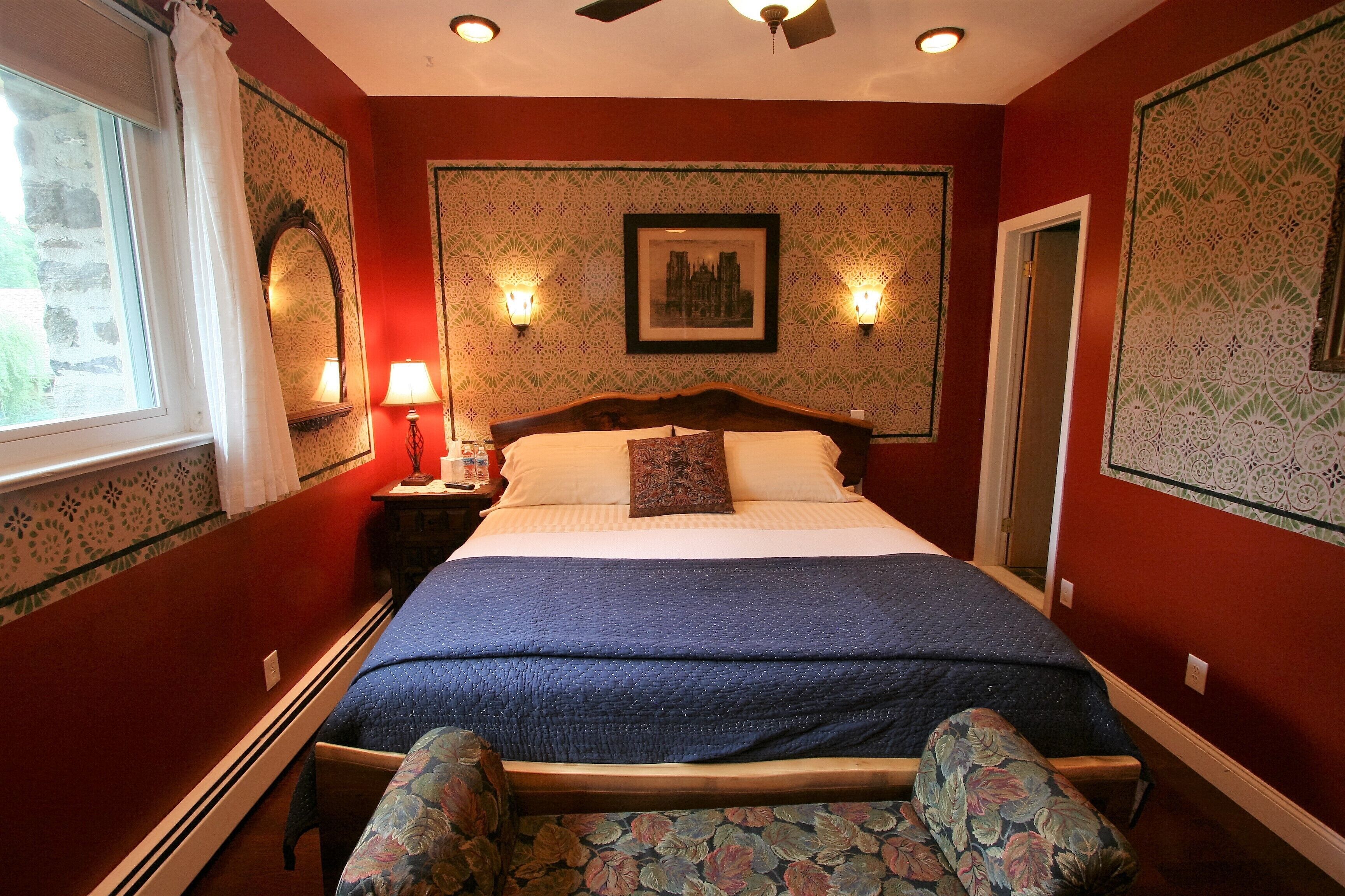A bedroom with a bed, nightstands, a window with curtains, a ceiling fan, and patterned wallpaper.