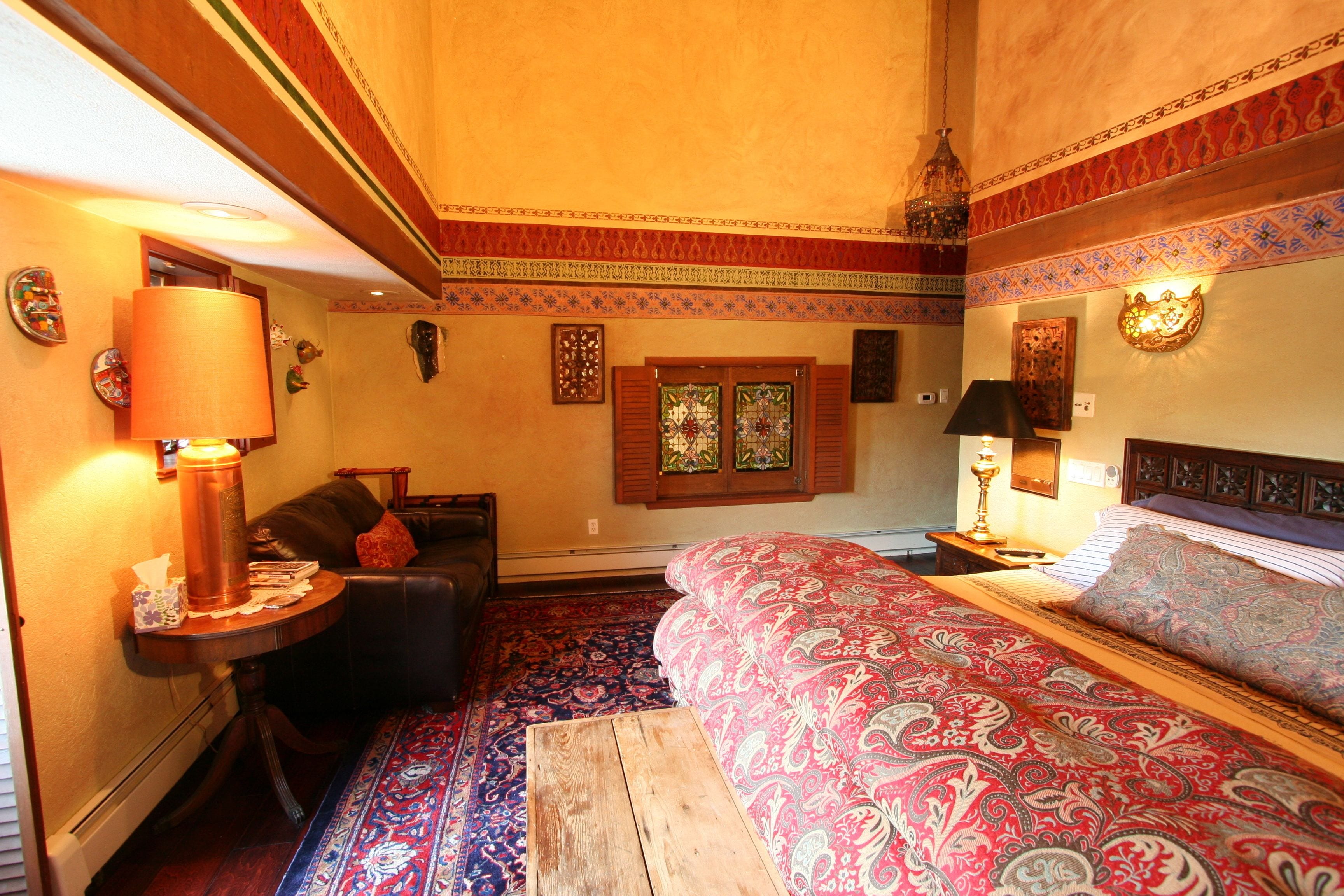 A room with a bed, a table with a lamp, a sofa, and a patterned rug.