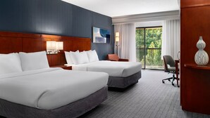 Une chambre d’hôtel avec deux lits, un bureau, une chaise, une lampe et une vue sur la verdure par la fenêtre.