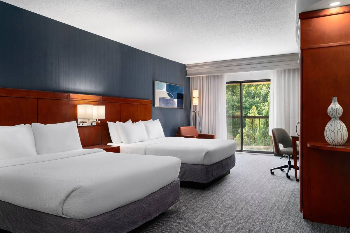 A hotel room with two beds, a desk, a chair, a lamp, and a view of greenery through the window.
