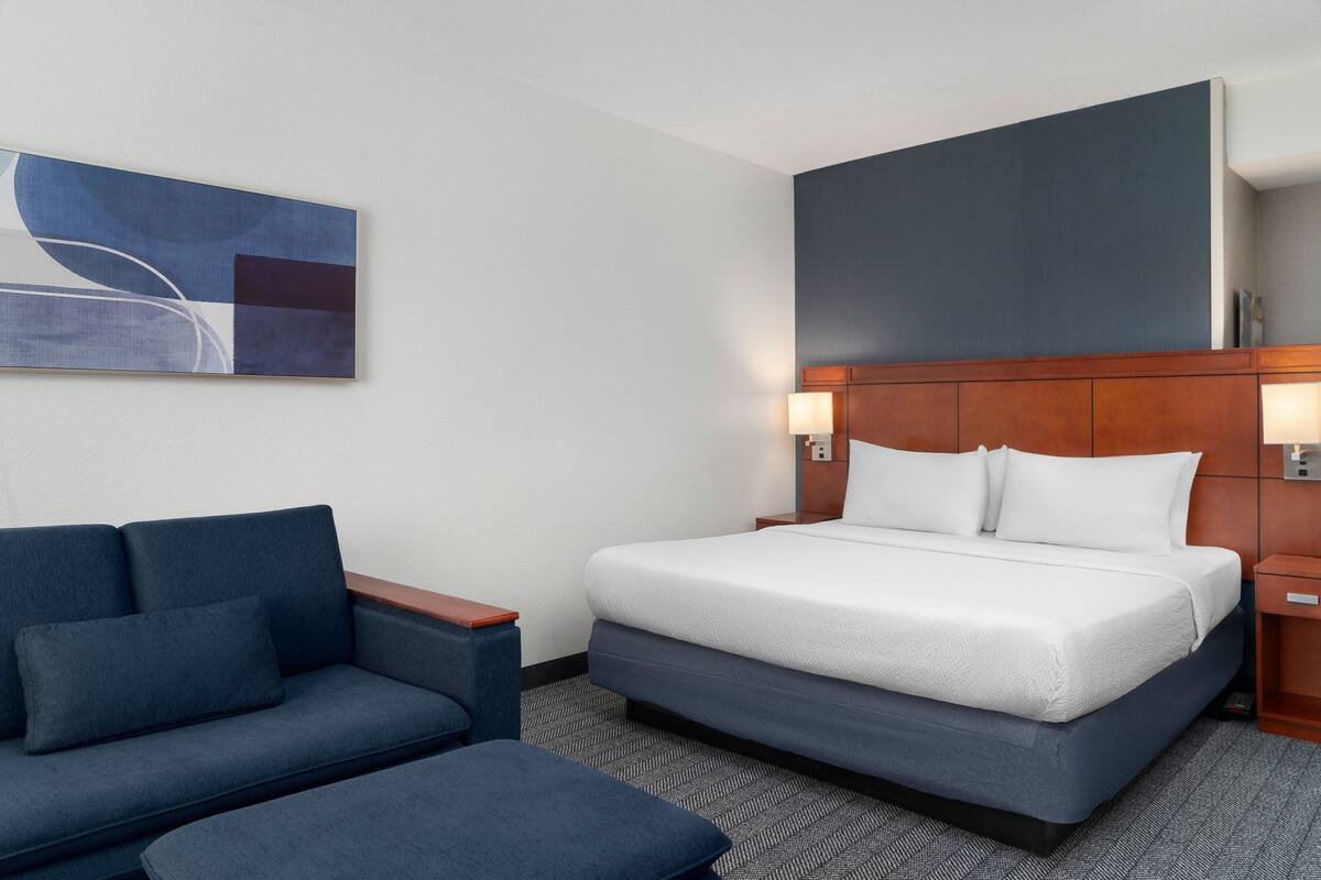 A hotel room with a bed, a sofa, a small table, and a wall-mounted artwork.