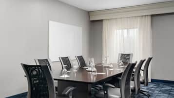A conference room with a long table, six chairs, a whiteboard, and a window with sheer curtains.