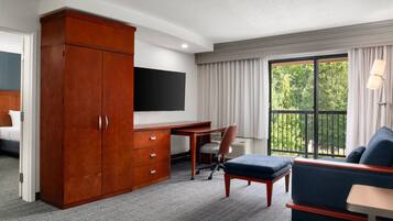 Habitación de hotel con armario de madera, escritorio con silla, sofá, televisor montado en la pared y balcón con cortinas.