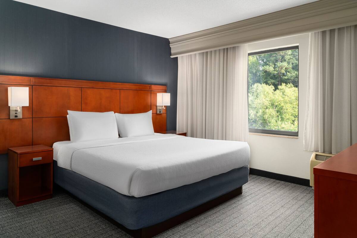 A hotel room with a large bed, wooden headboard, two bedside lamps, a nightstand, and a window with curtains.