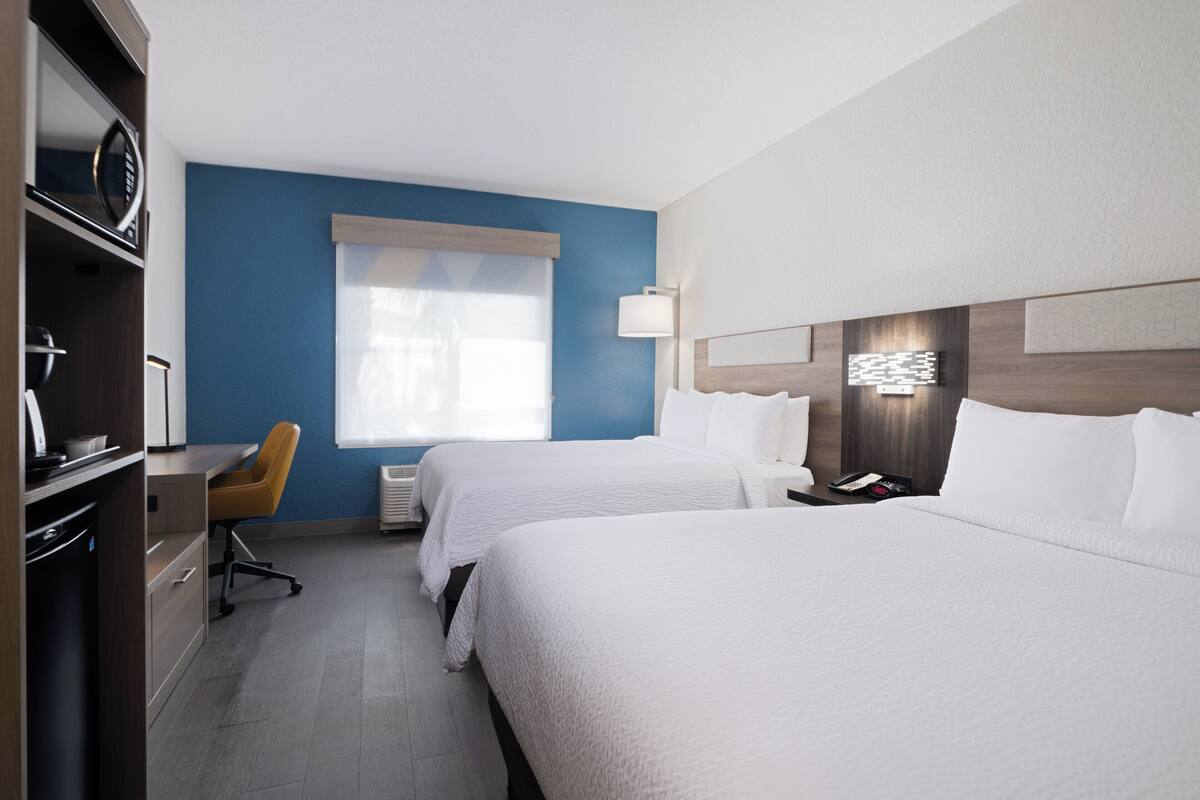 A hotel room with two beds, a desk, a chair, and a large window with blinds.
