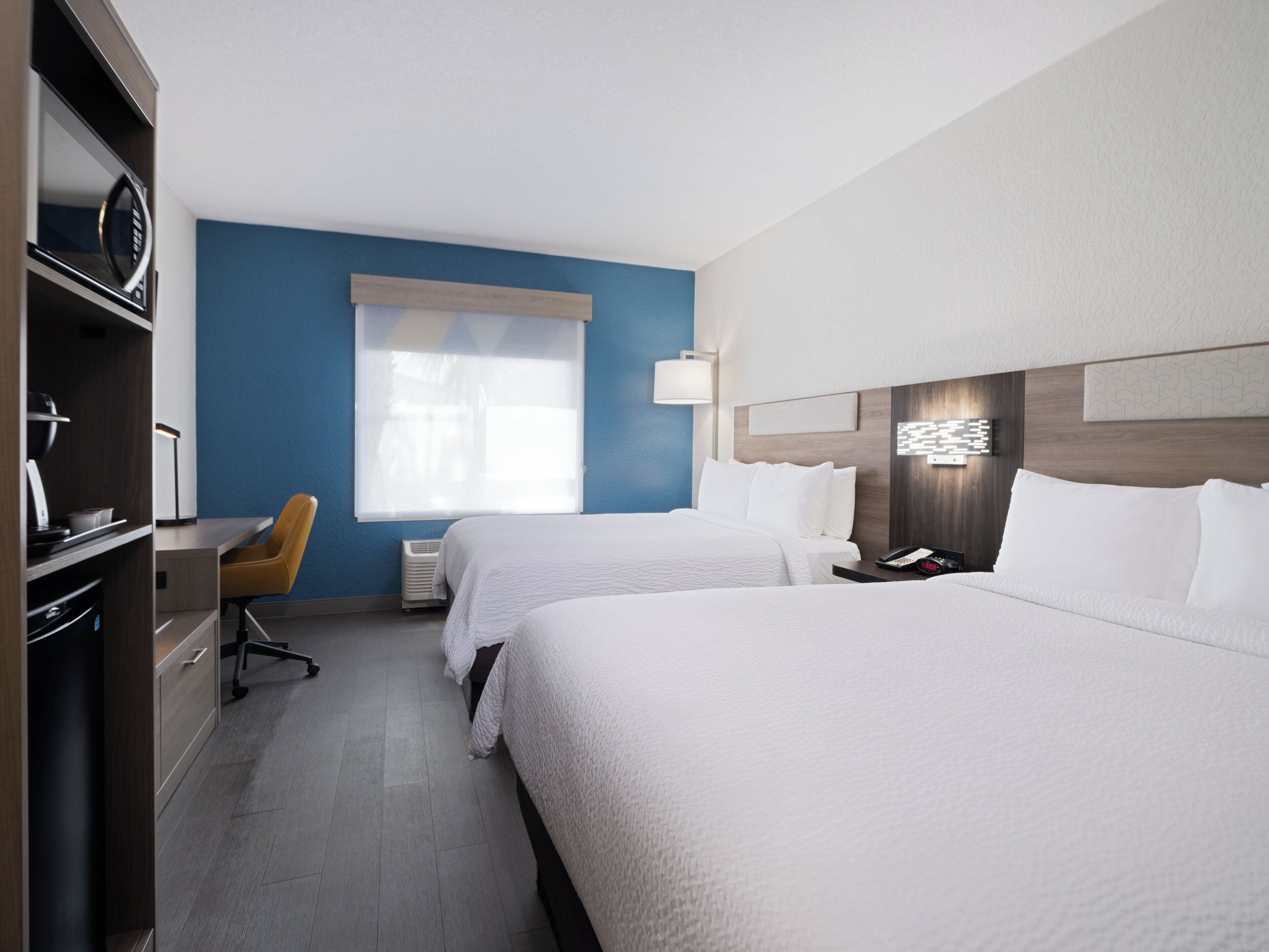 A hotel room with two beds, a desk, a chair, and a large window with blinds.