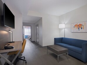 Una habitación de hotel moderna con escritorio, silla, sofá y televisor de pantalla plana montado en la pared.