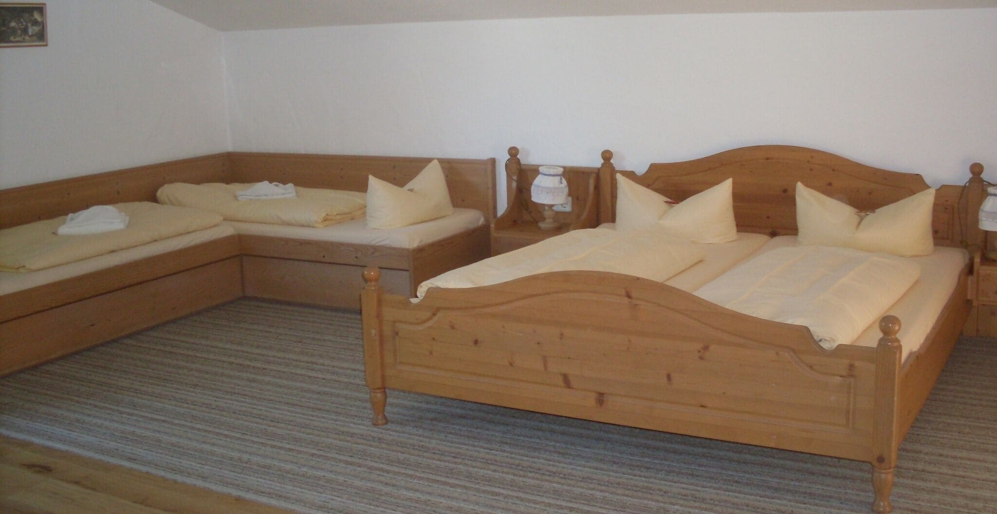 A room with two wooden beds, each with a wooden headboard and a bedside table. The room has a carpeted floor and plain white walls.