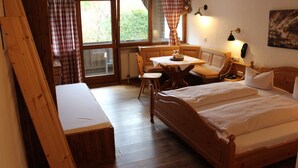 Un lit en bois, accompagné d’un banc, d’une petite table et d’une chaise, dans une pièce dotée de planchers en bois et de grandes fenêtres.