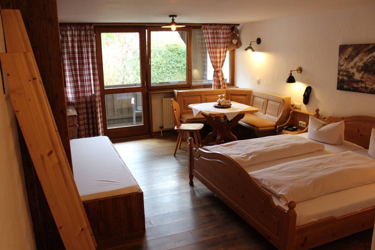 Un lit en bois, accompagné d’un banc, d’une petite table et d’une chaise, dans une pièce dotée de planchers en bois et de grandes fenêtres.