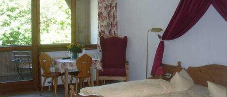 A bedroom with a double bed, a chair, a table with a vase of flowers, and a window with curtains.