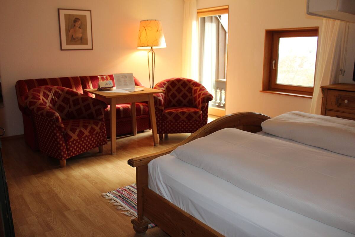 Une chambre d’hôtel avec un lit, un bureau doté d’une lampe, deux fauteuils à motifs, une petite table et une fenêtre avec des rideaux.