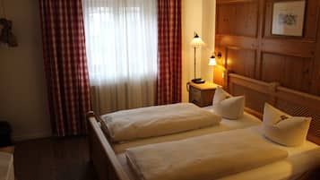 A hotel room with two beds, a wooden headboard, a nightstand with a lamp, and a window with red and white curtains.