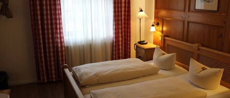 A hotel room with two beds, a wooden headboard, a nightstand with a lamp, and a window with red and white curtains.