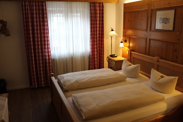 A hotel room with two beds, a wooden headboard, a nightstand with a lamp, and a window with red and white curtains.