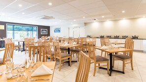 A spacious dining area with multiple tables and chairs, a kitchen area with buffet setup, and a large window providing natural light.