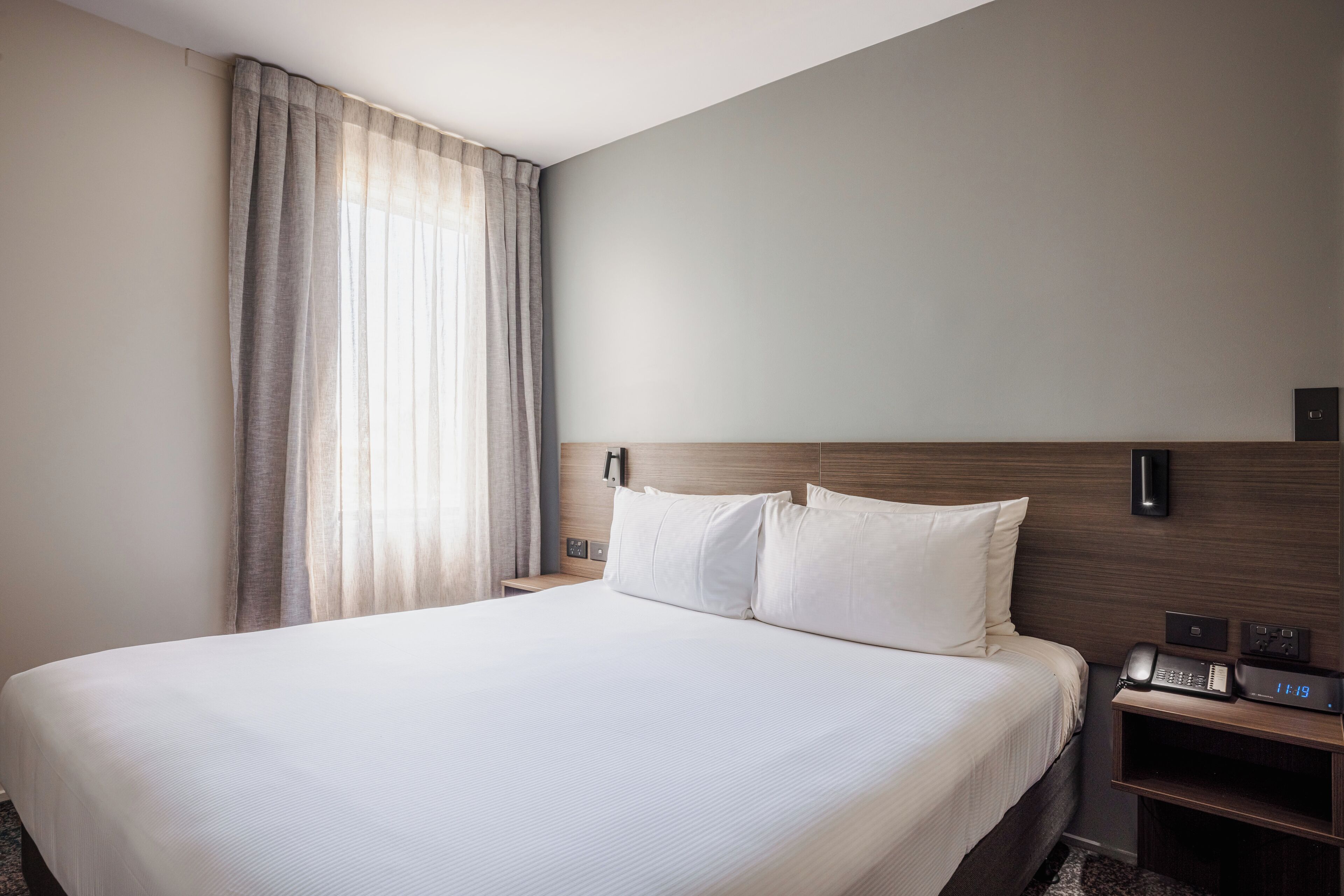 A hotel room with a large bed, a bedside table with a phone and a digital clock, and a window with curtains.