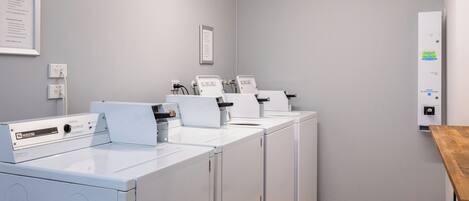 A laundry room with multiple white washing machines and dryers, a wooden table, and a wall-mounted electrical panel.
