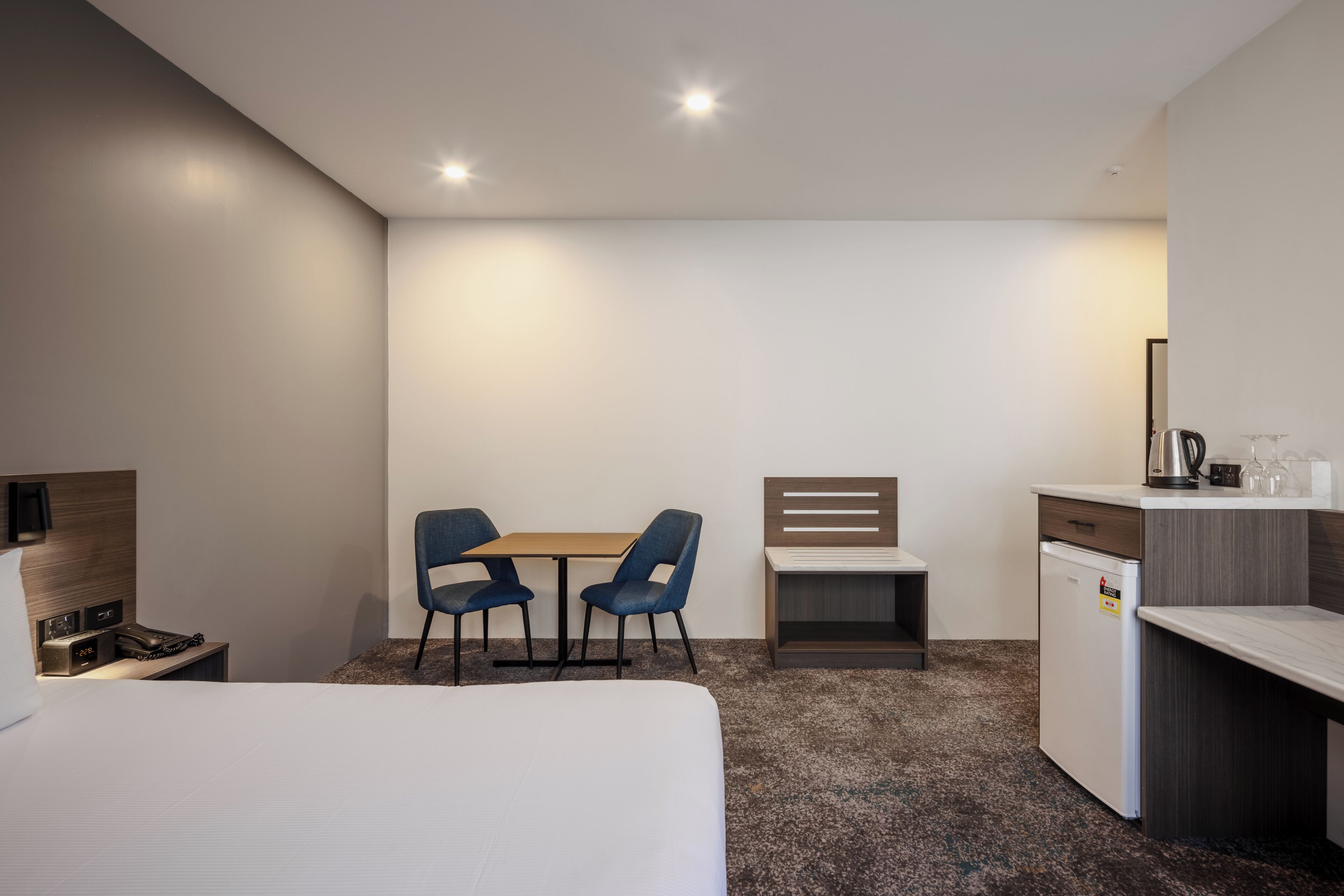 Quarto de hotel com cama, escrivaninha, duas cadeiras, pequeno espaço de cozinha e mesa de cabeceira com luminária.