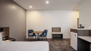 Quarto de hotel com cama, escrivaninha, duas cadeiras, pequeno espaço de cozinha e mesa de cabeceira com luminária.