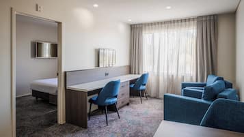A hotel room with a bed, a desk with two chairs, a television, and a large window with curtains.