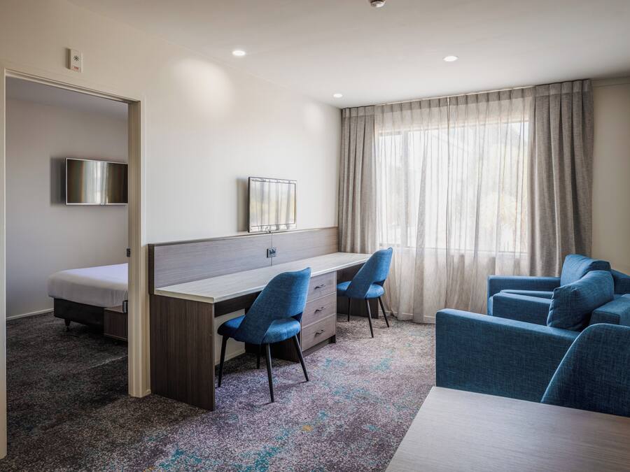 A hotel room with a bed, a desk with two chairs, a television, and a large window with curtains.