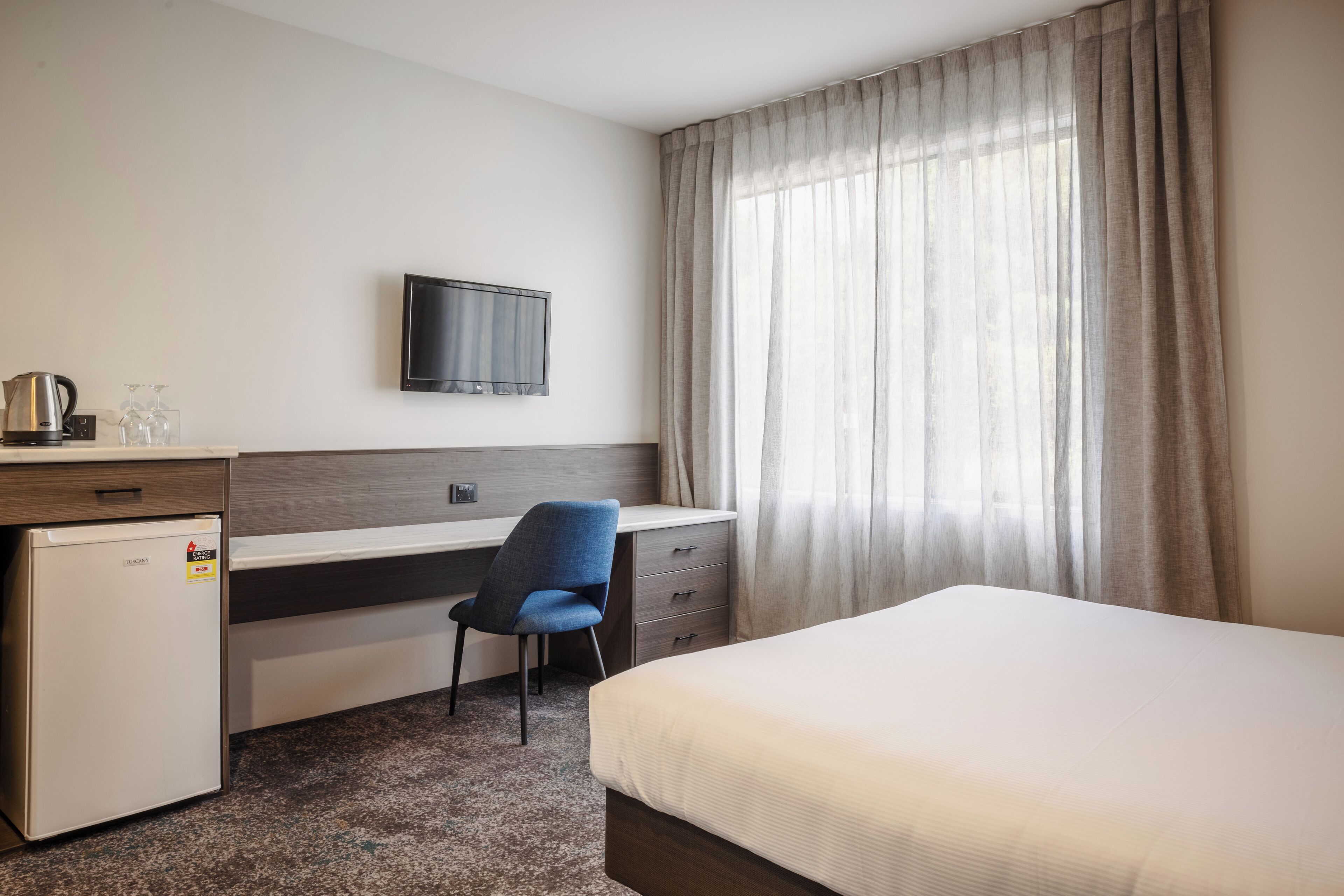 A hotel room with a bed, a desk with a chair, a television, and a small refrigerator.