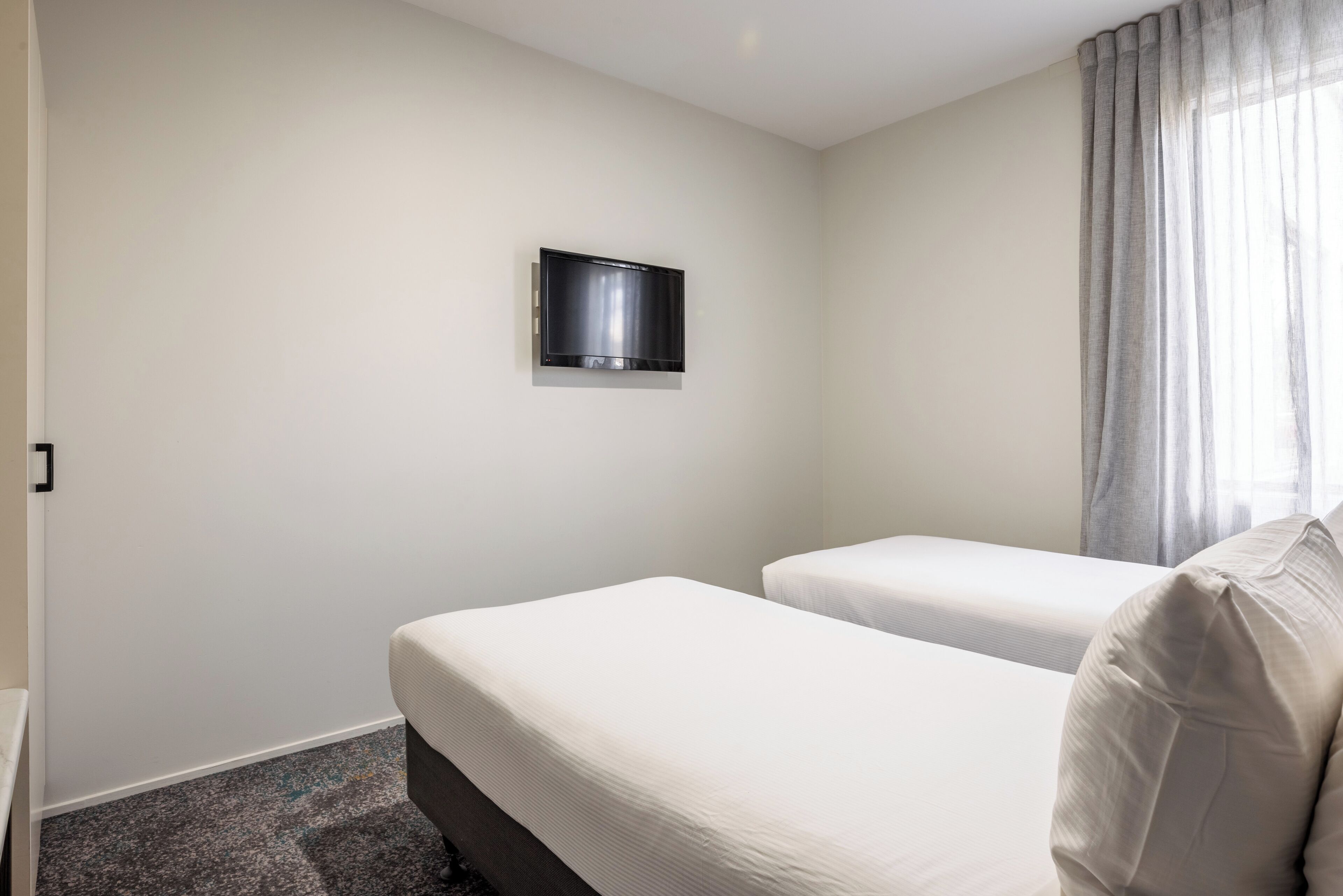 Habitación de hotel con dos camas individuales, un televisor de pantalla plana montado en la pared y una ventana con cortinas.