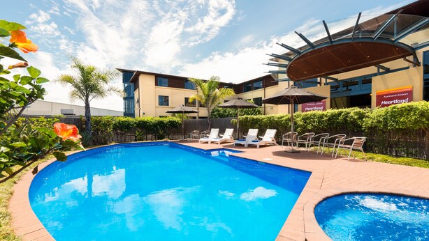 Una piscina con sillas de descanso y sombrillas, rodeada de un jardín ajardinado y un edificio con señalización para el Aeropuerto Heartland de Auckland.