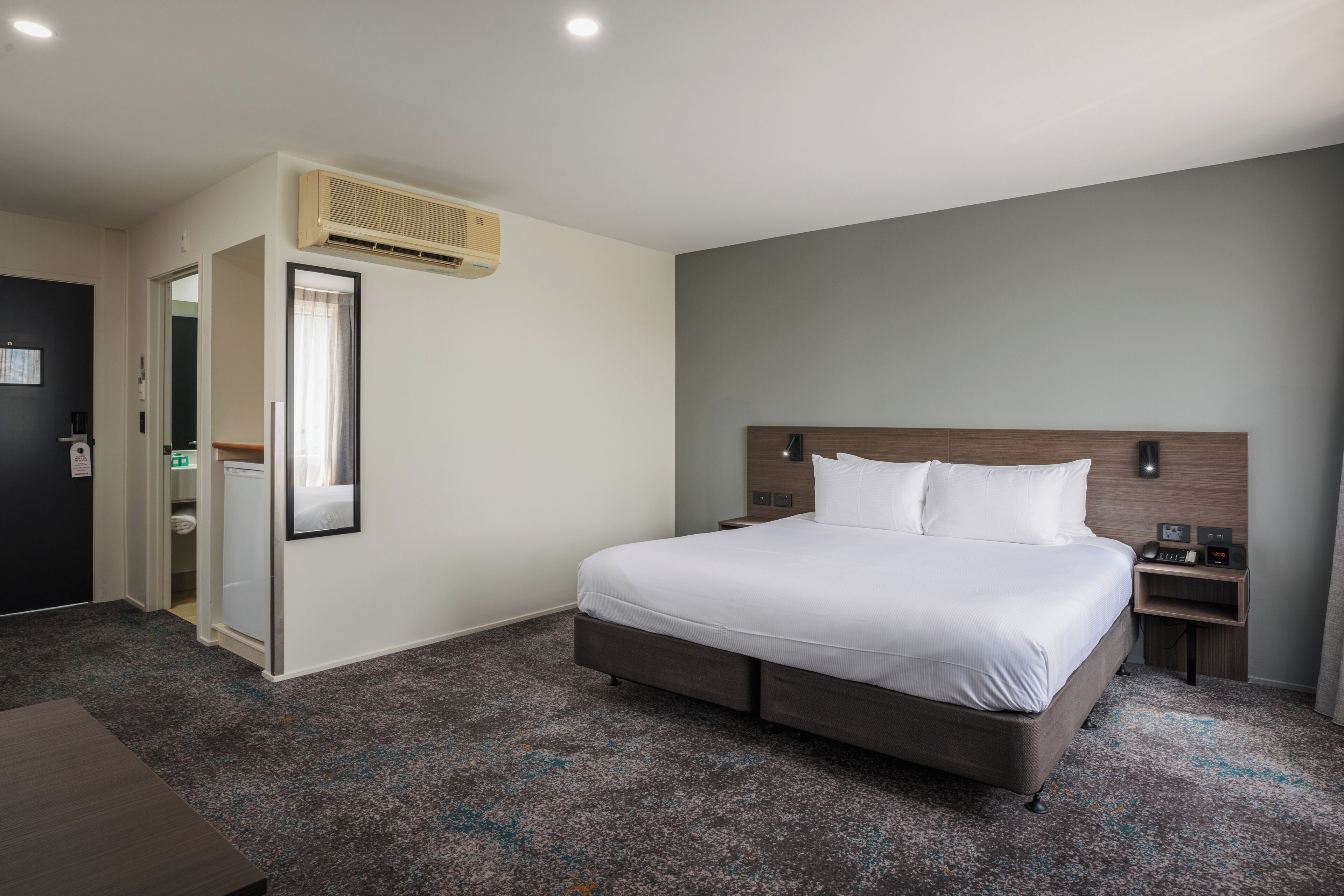 A hotel room with a large bed, a bedside table, a nightstand, a wardrobe, and an air conditioning unit.