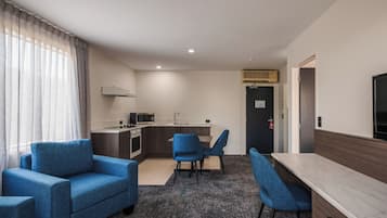 A modern hotel room with a kitchenette, a dining area with blue chairs, a sofa, and a large window with sheer curtains.