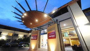 Una entrada de hotel moderna con una estructura distintiva con forma de sol sobre las puertas, señalización clara para el Auckland Airport Heartland Hotels, y un área de estacionamiento visible con coches.