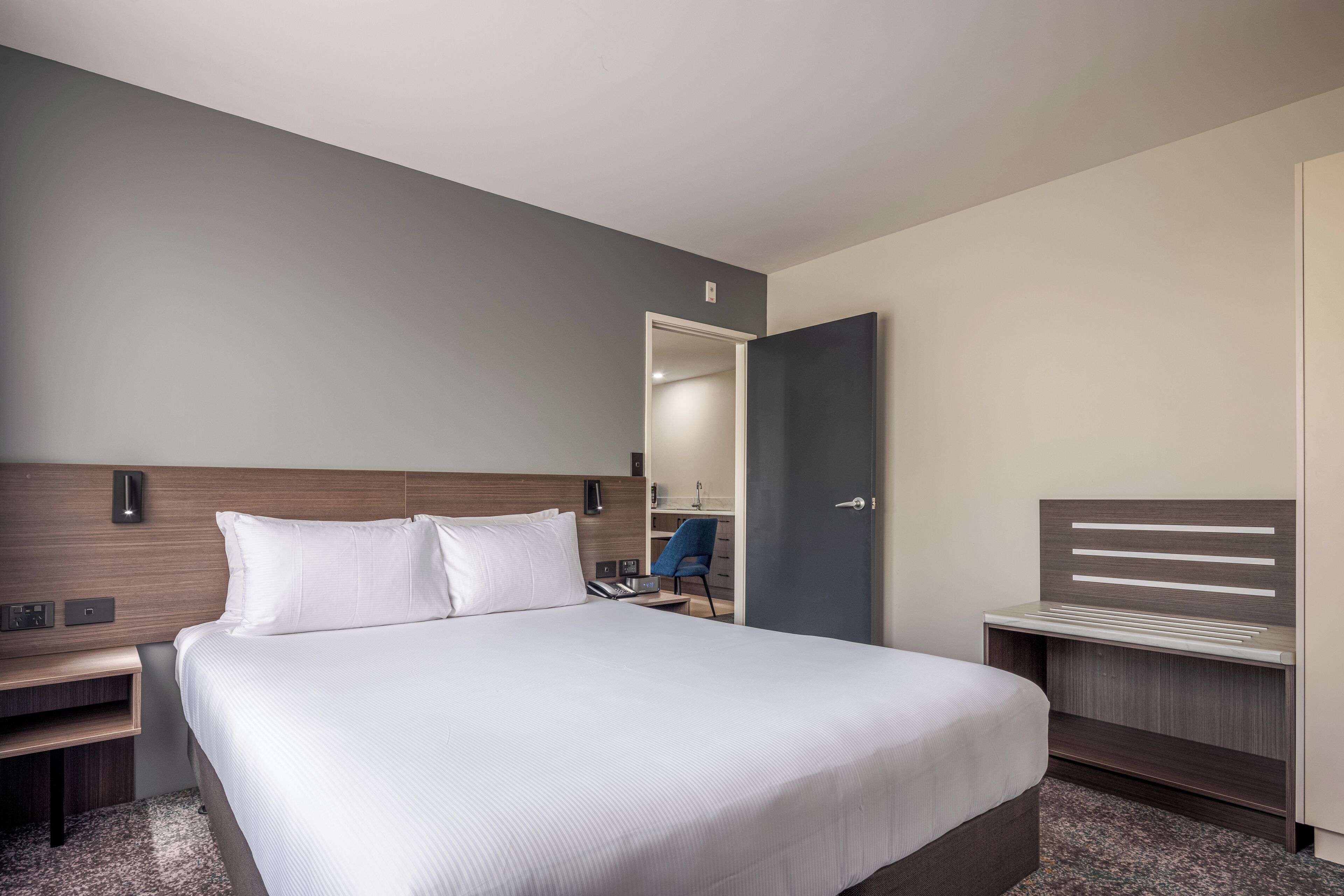 A modern hotel room with a large bed, a bedside table, a chair, and a wardrobe.