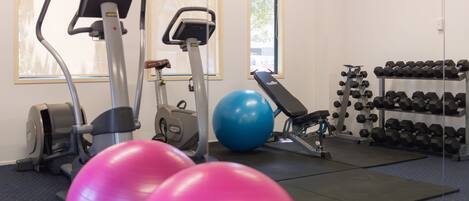 Un gimnasio con equipo de ejercicio, una pelota de ejercicio azul, pelotas de ejercicio rosas y un estante con mancuernas.