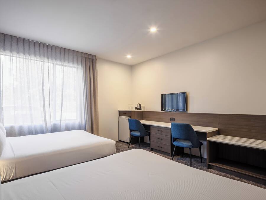 Une chambre d’hôtel avec deux lits, un bureau, deux chaises, un petit réfrigérateur et une télévision.