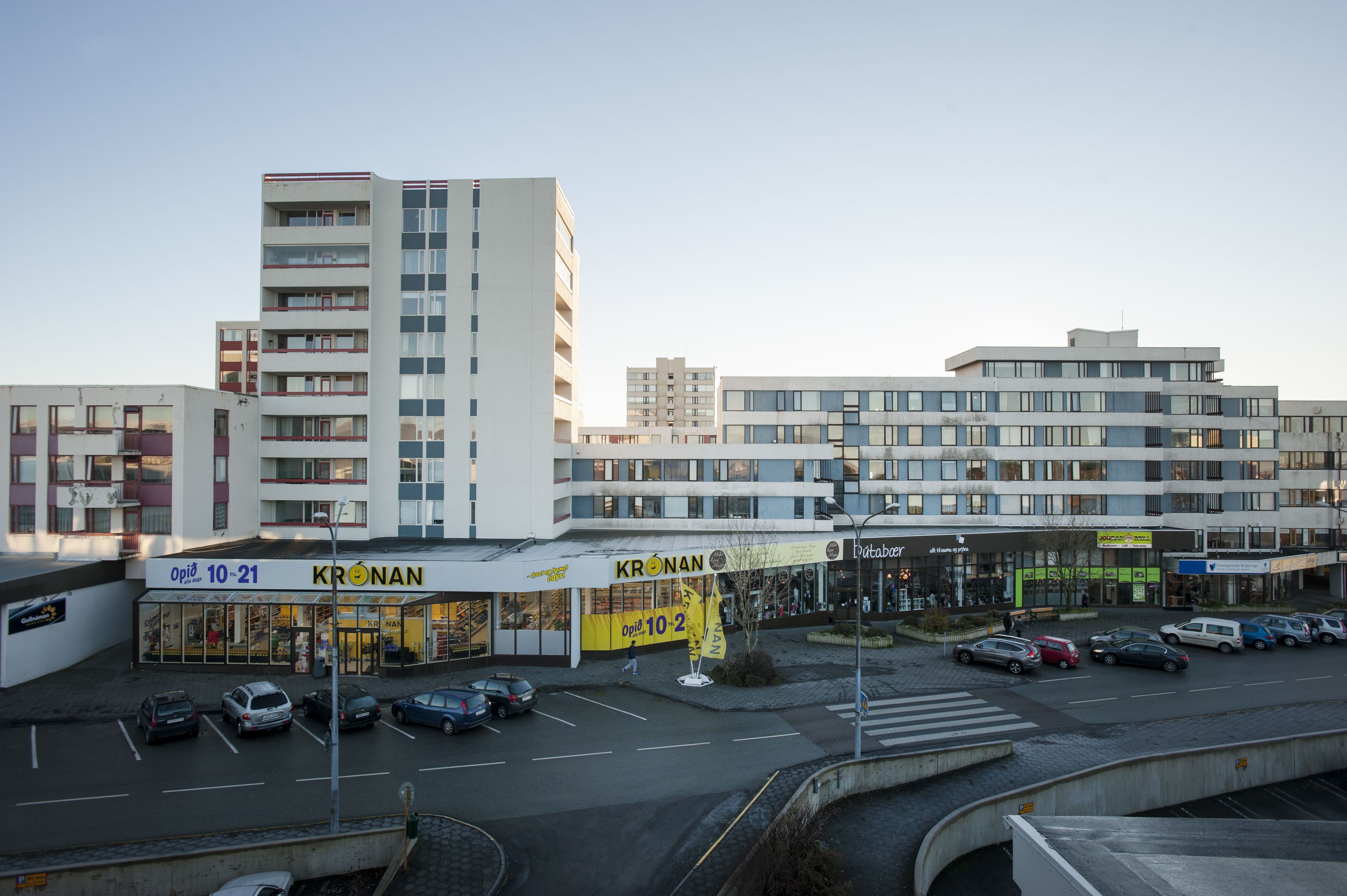 A multi-story building with a prominent 'KRONAN' sign, a parking lot with several cars, and a clear sky.