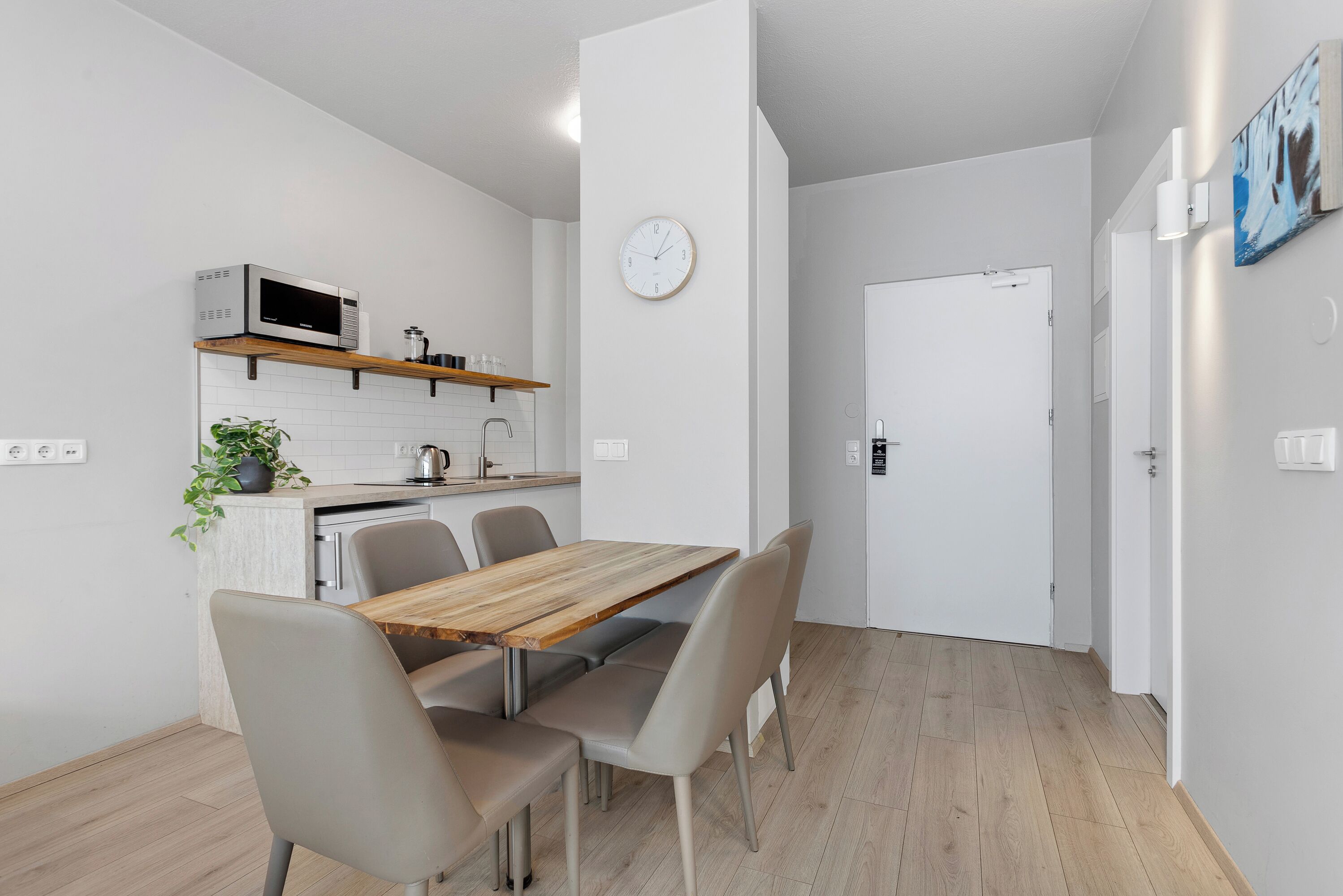Una cocina moderna con microondas, lavaplatos y encimera de madera. Se aprecia una mesa de comedor con sillas y un reloj en la pared.