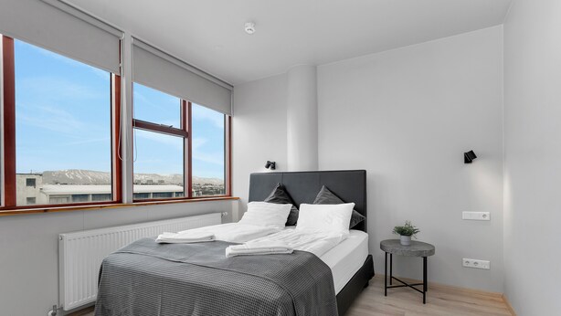 A modern bedroom with a large window, a bed with a grey and white checkered bedspread, a small bedside table with a plant, and a radiator. - Iceland Comfort Apartments by Heimaleiga (Kópavogur)