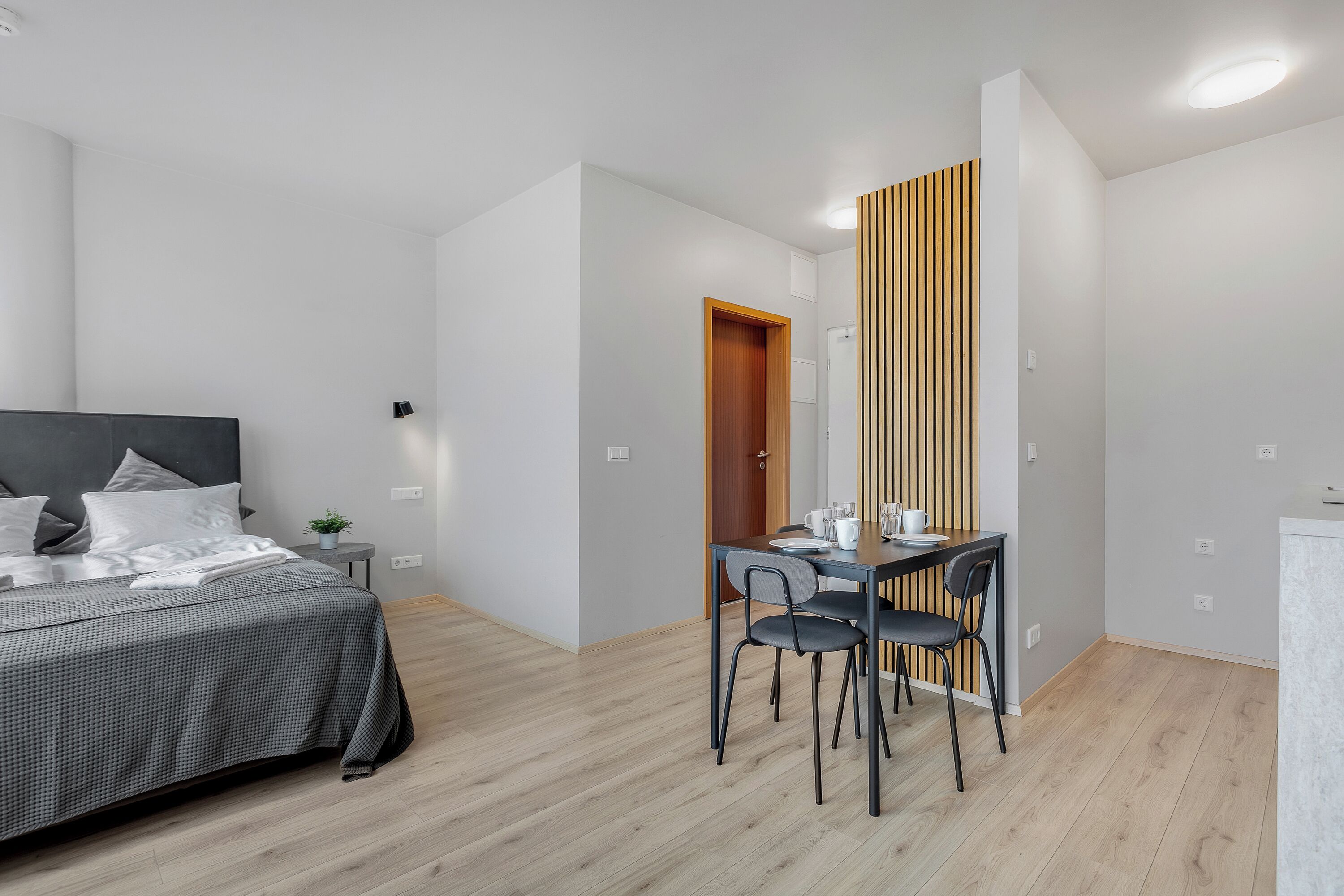 A modern bedroom with a bed, bedside table, and a small plant. There is a separate dining area with a table and chairs.