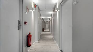 Un pasillo largo y estrecho con paredes blancas y suelo de madera clara. Se observan extintores montados en las paredes y un detector de incendios en el lado izquierdo.