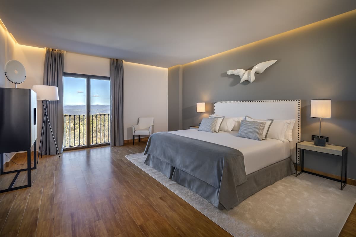 A modern bedroom with a large bed, a balcony view, and a desk with a lamp.