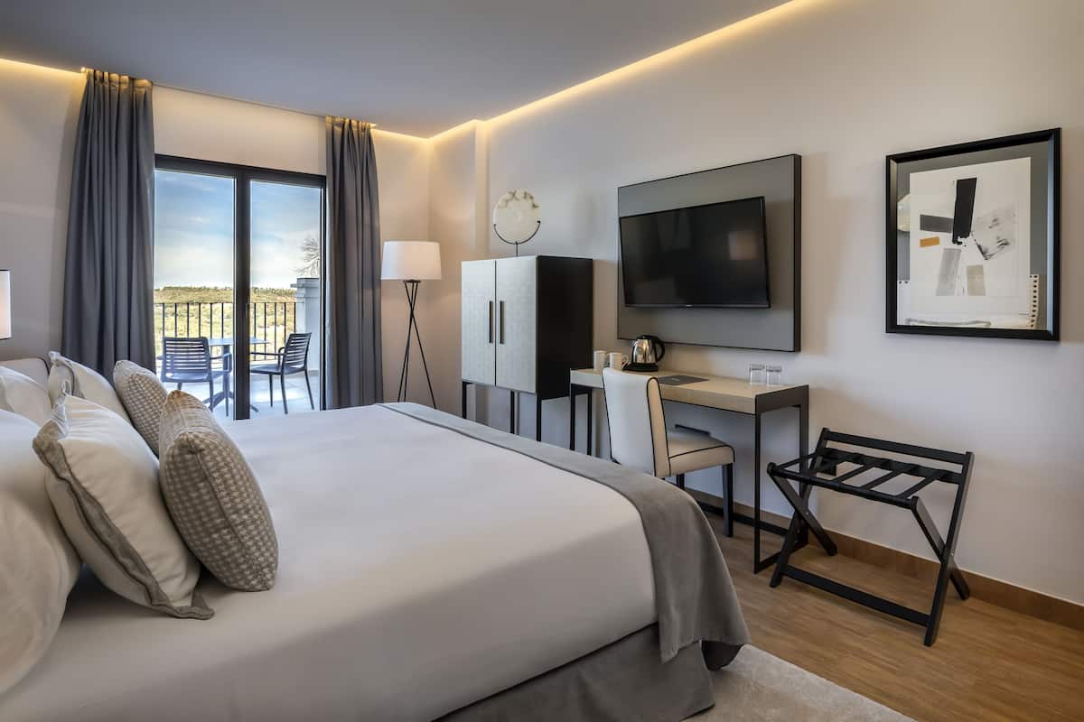 A modern hotel room with a large bed, a desk, a TV, and a balcony with outdoor seating.