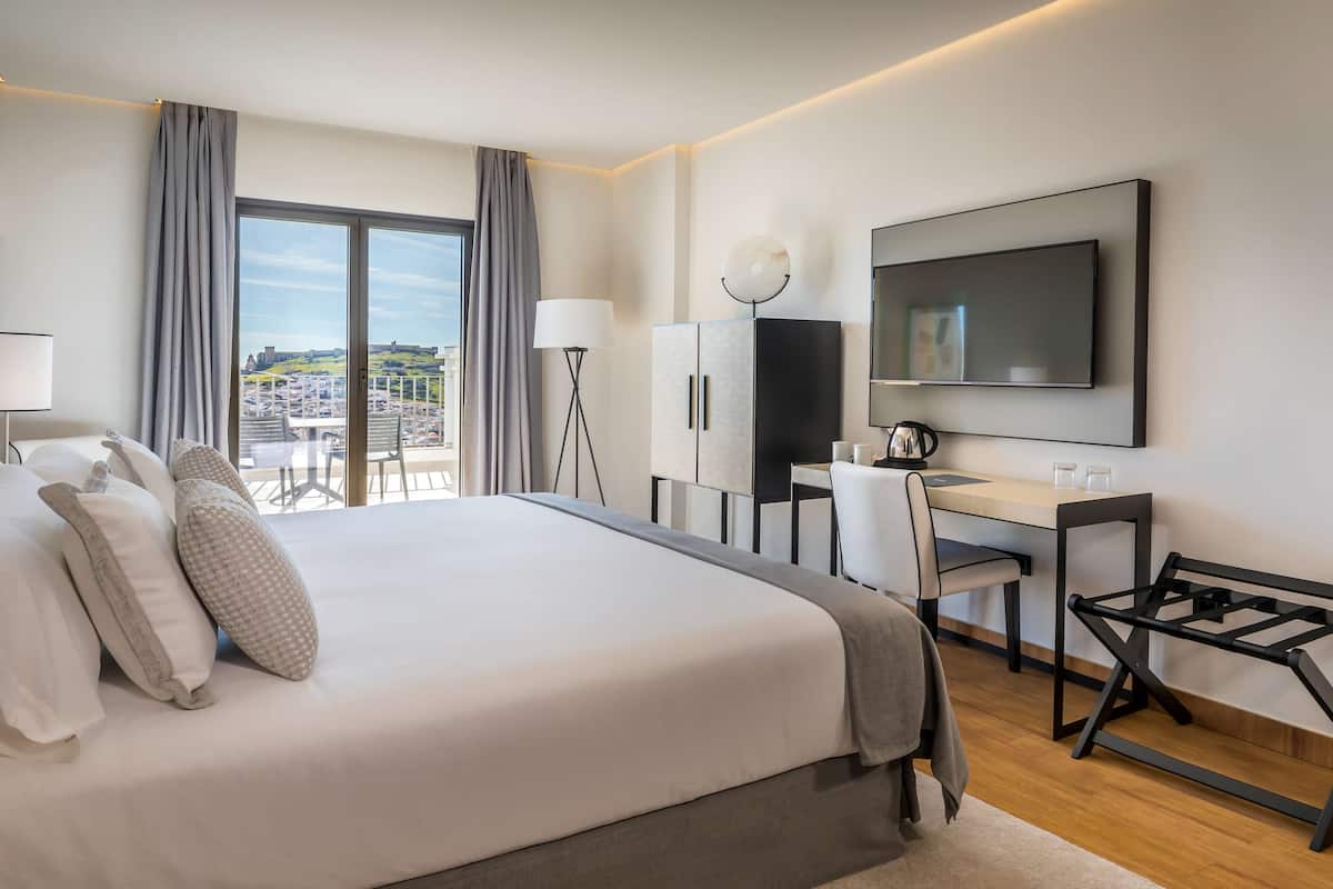 A modern hotel room with a large bed, a desk, a TV, and a view of the city.