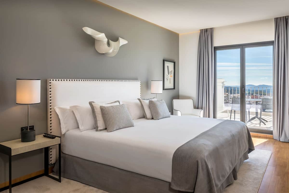 A modern hotel room with a large bed, a bedside table with a lamp, a wall-mounted light fixture, and a view of the city through large windows.
