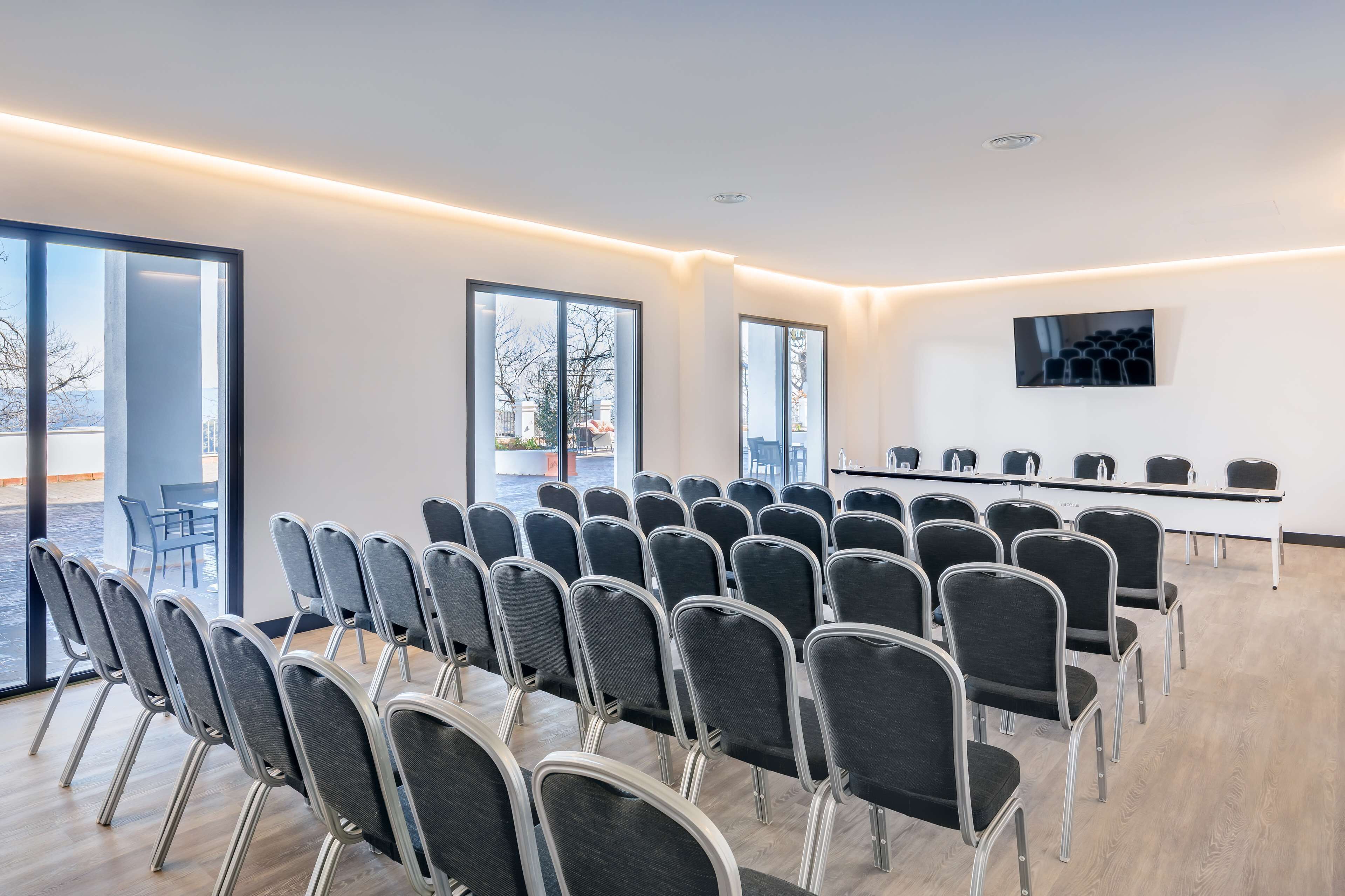 Una sala de conferencias con filas de sillas, una mesa larga y una pantalla de televisión en la pared.