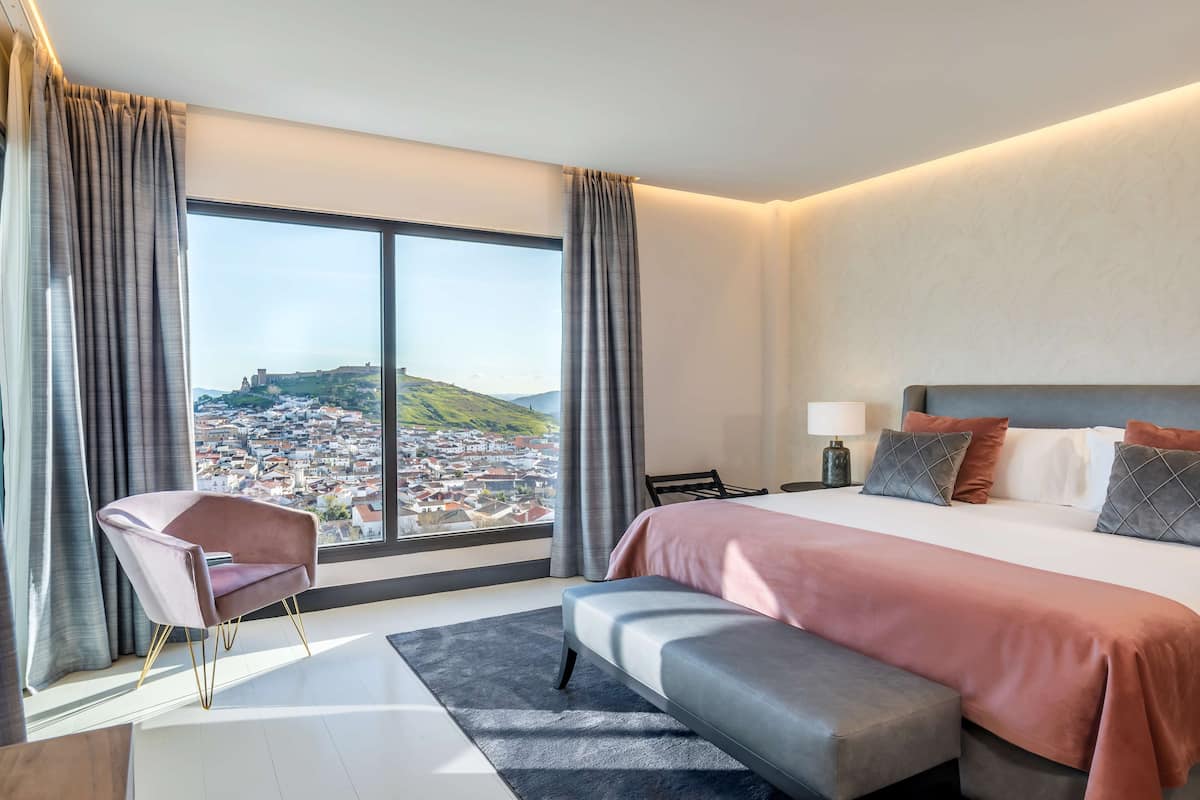 A modern hotel room with a large bed, a view of a cityscape, a chair, and a bench.