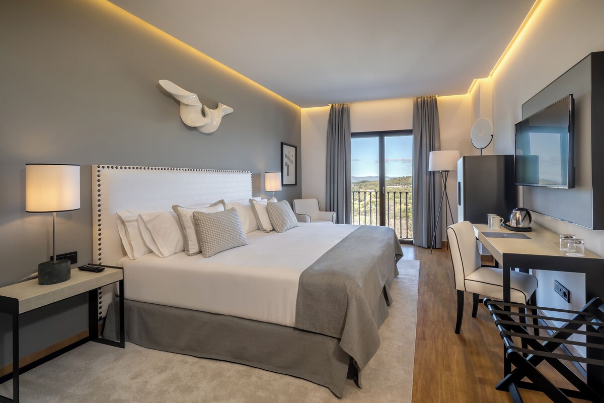 A modern hotel room with a large bed, a desk with a chair, a TV, and a balcony with a view.